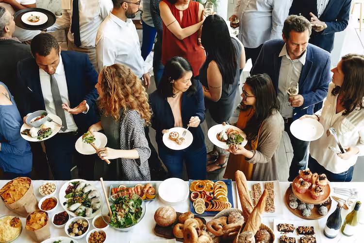A group of people dressed in business casual clothing. The photo is taken from above and you can see that the corporate event is catered, everyone is eating and talking.